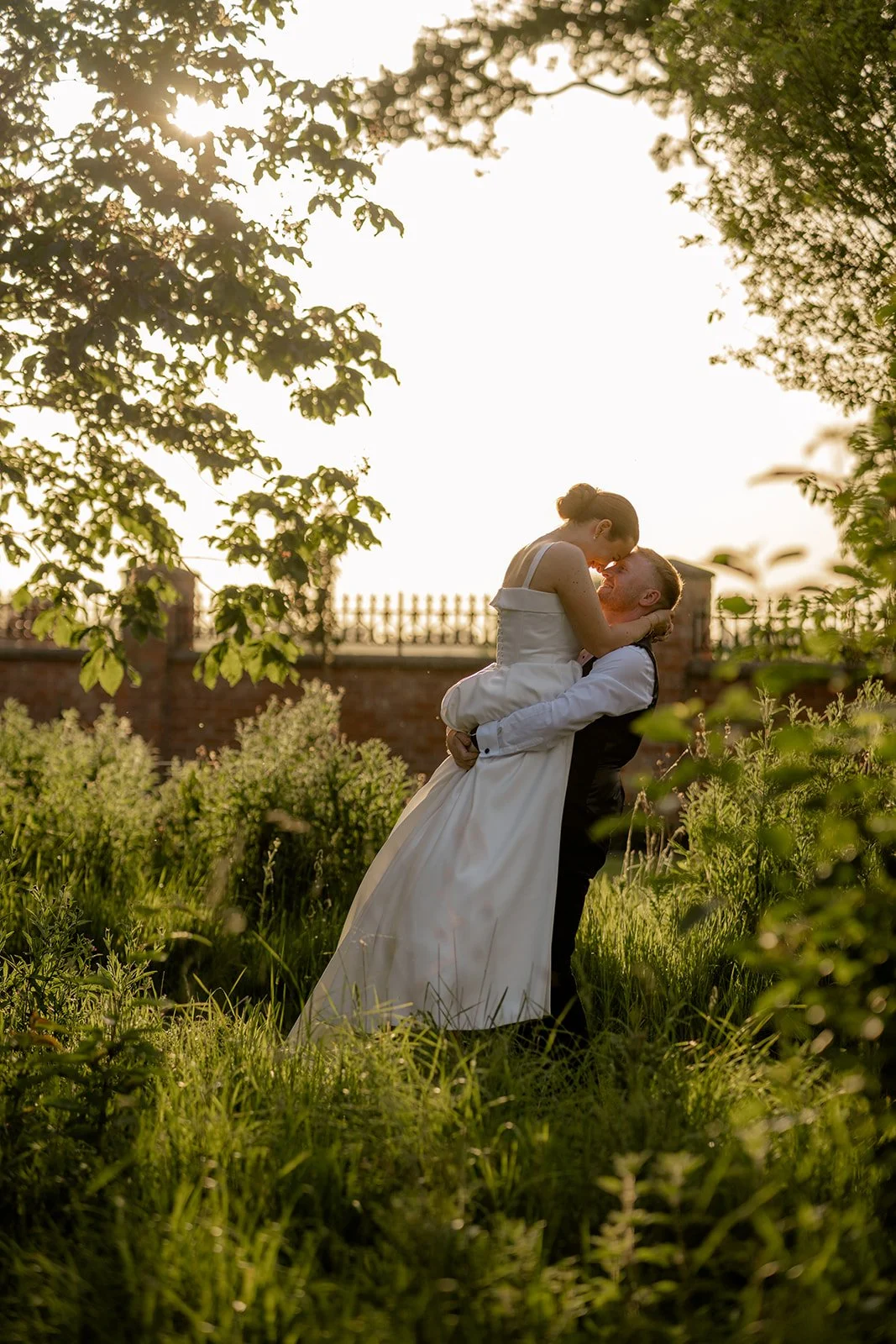 Golden hour wedding portrait of Lydia & Kieran laughing as the groom lifts the bride in long grass at Hencote Estate in Shropshire, UK, surrounded by greenery, warm summer light and relaxed countryside wedding photography moments together.