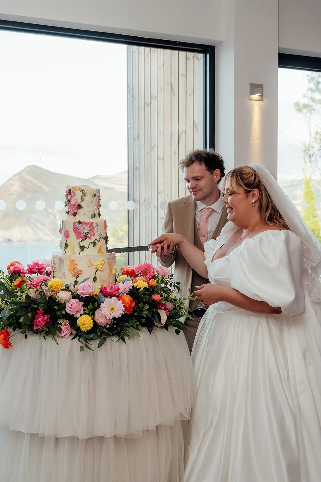Holly & Alex cut their colourful floral wedding cake beside bright flowers and coastal views at Sandy Cove Hotel in North Devon, photographed by Just James Weddings.