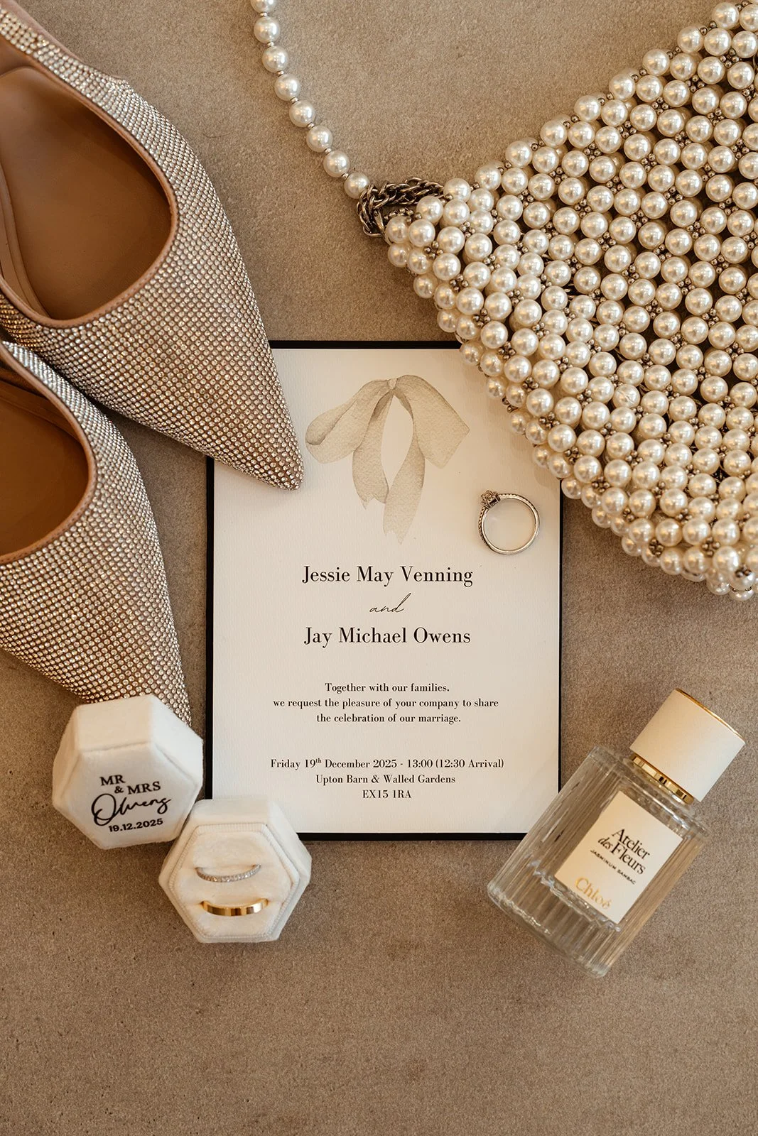 Wedding flat lay featuring Jessie & Jay’s invitation suite, pearl bag, bridal heels, rings, and perfume, styled together at Upton Barn & Walled Garden in Devon, UK. Neutral tones and refined details reflect a modern, elegant wedding aesthetic.