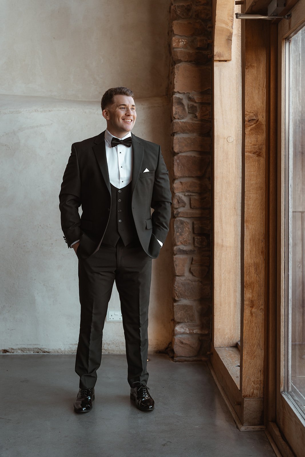 Jay standing by a window in his black tuxedo during wedding morning preparations at Upton Barn & Walled Garden in Devon, UK. Natural light and textured stone walls frame a relaxed, confident portrait of Jay ahead of marrying Jessie.