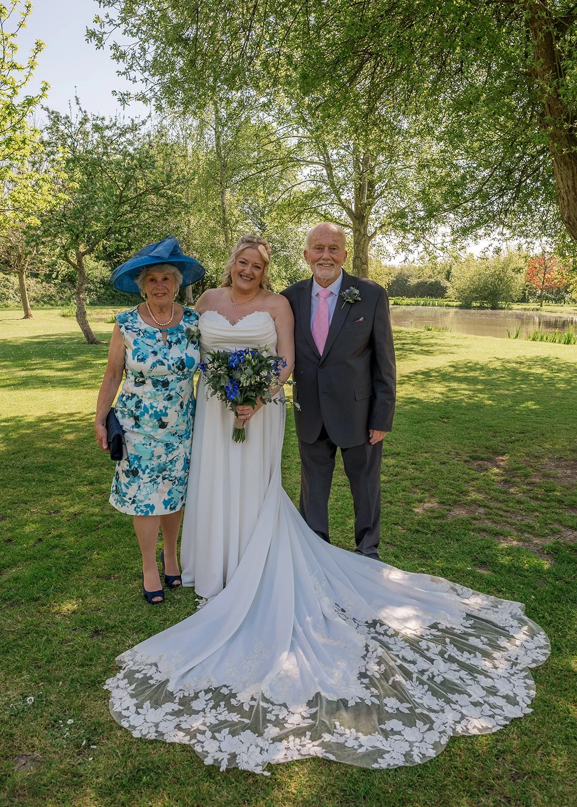 Debbie poses with her parents beside the lake at Yarlington Barn in Somerset, holding her blue wedding bouquet as her lace train spreads across the grass for a sunny family portrait on Debbie and Nigel’s countryside wedding.
