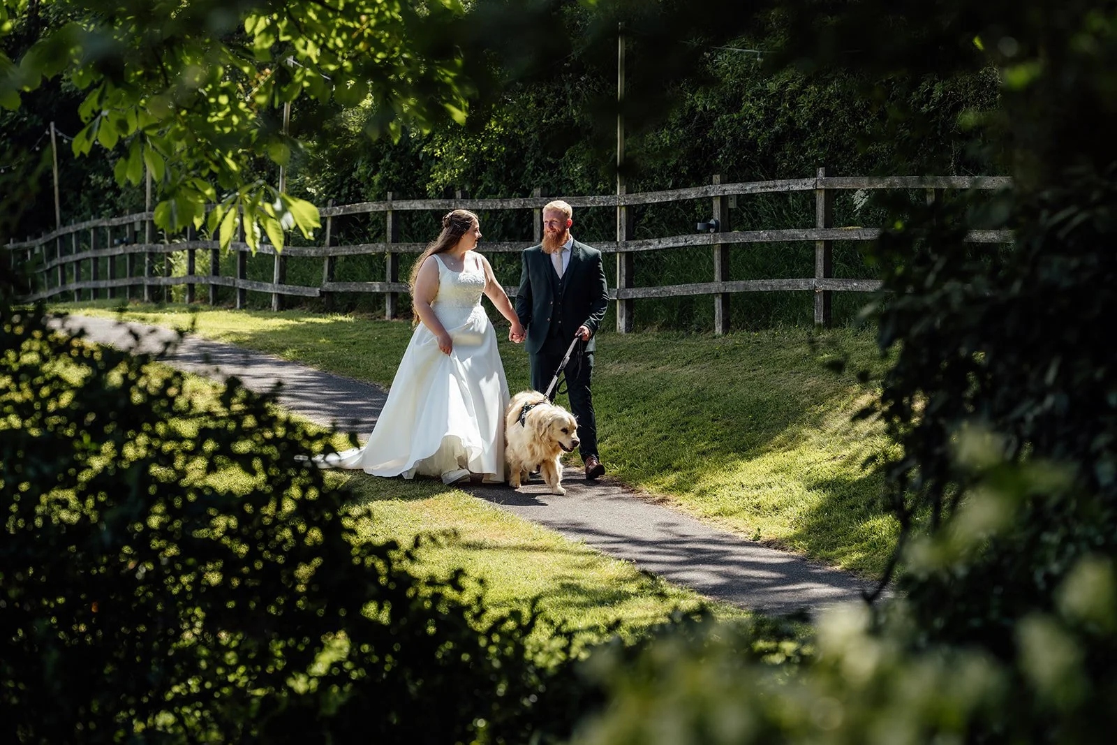 Romantic countryside walk for Louise & Jon at Wick Farm Bath in Somerset, UK, with the couple holding hands as they stroll with their golden retriever along a sunny garden path, framed by leafy trees, rustic fencing, and summer light.