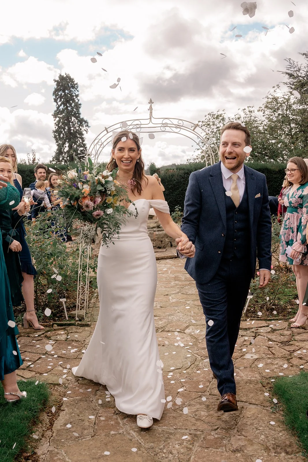 Lilly & Lloyd walking hand in hand through confetti at Broadfield Court in Herefordshire, photographed by Just James Weddings for joyful documentary wedding photography.