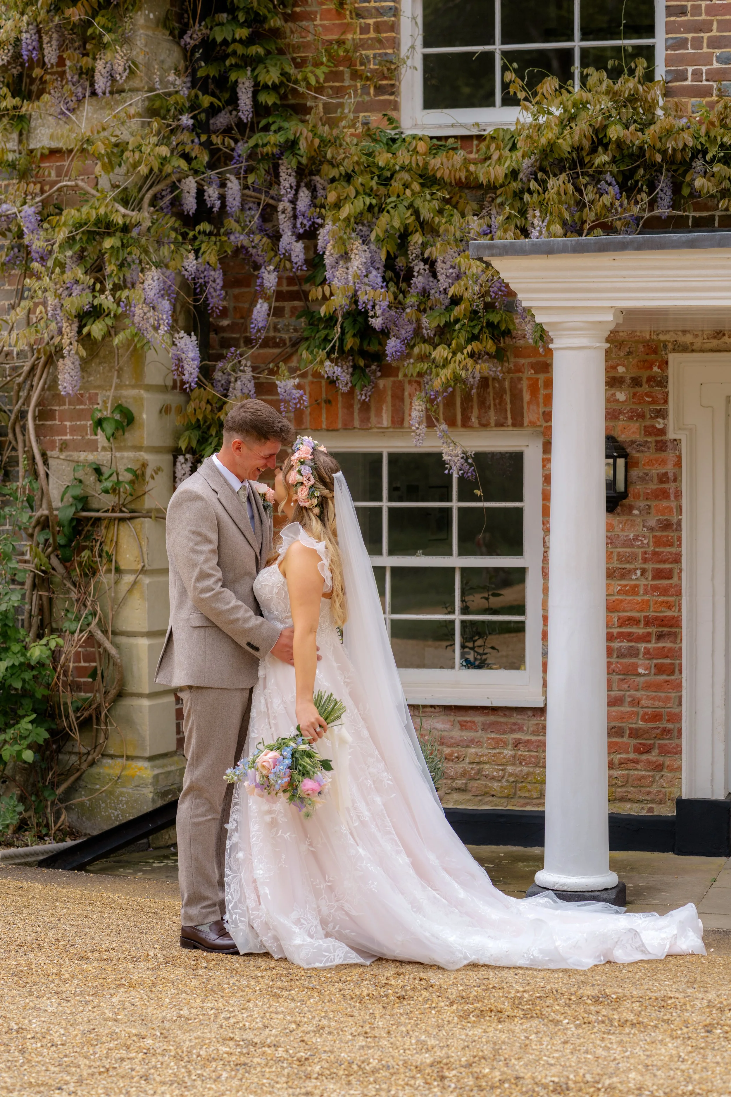 Jaye & Josh’s wedding at Syrencot in Wiltshire, captured in a romantic couple portrait outside the brick manor house, with wisteria, white columns, a flowing veil, pastel bouquet and the newlyweds sharing a quiet kiss together in the courtyard.