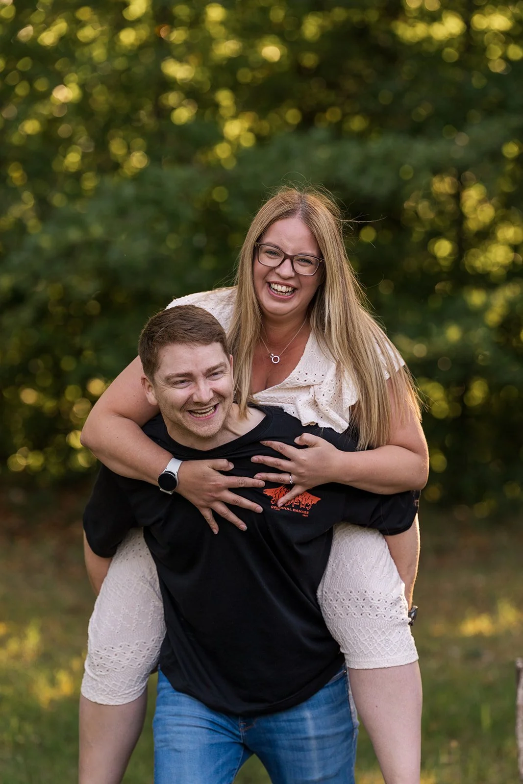 Chloe laughing on Rob’s back during a playful Somerset engagement photoshoot, with both smiling at the camera in soft evening light against a leafy green backdrop, captured naturally by Just James Weddings before their upcoming wedding day.