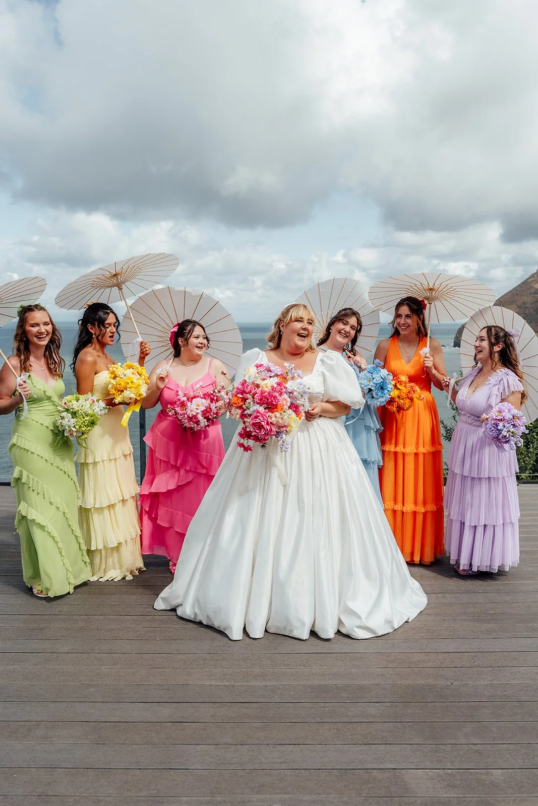 Holly laughs with her bridesmaids in colourful dresses as they hold white parasols and bright bouquets overlooking the coast at Sandy Cove Hotel in North Devon, photographed by Just James Weddings.