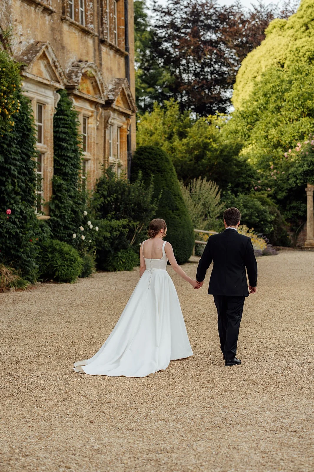 Rachel & Tom walk hand in hand along a gravel path beside the historic house and gardens at Brympton House in Somerset, photographed by Just James Weddings.
