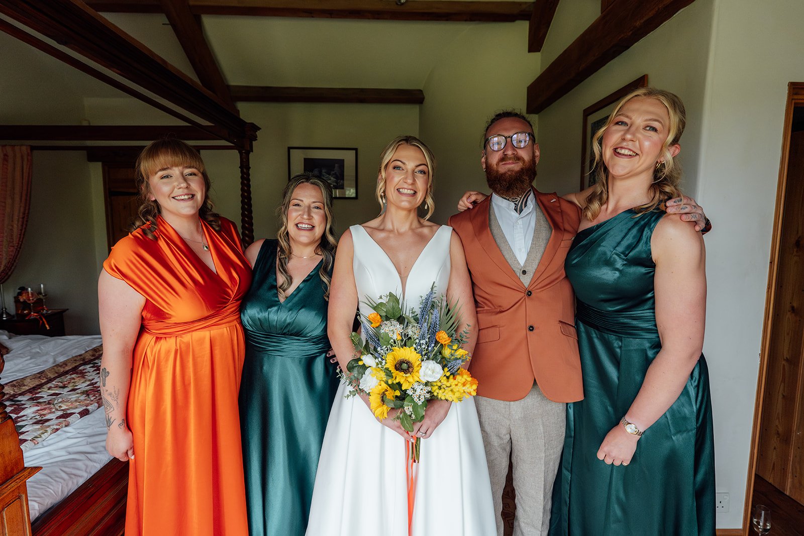 Joyful wedding party portrait with Hannah and her closest people at Grays Barn in Devon, UK, featuring emerald green and orange outfits, a sunflower bouquet and relaxed smiles during bridal preparations, photographed by Just James Weddings.