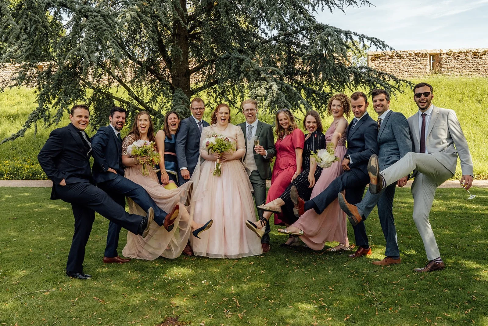 Charlotte & Jack wedding party portrait at Bishop’s Palace in Somerset, UK, with the couple standing among friends in suits and colourful dresses beneath a large tree, laughing as guests kick up their shoes on the sunny lawn after the ceremony.