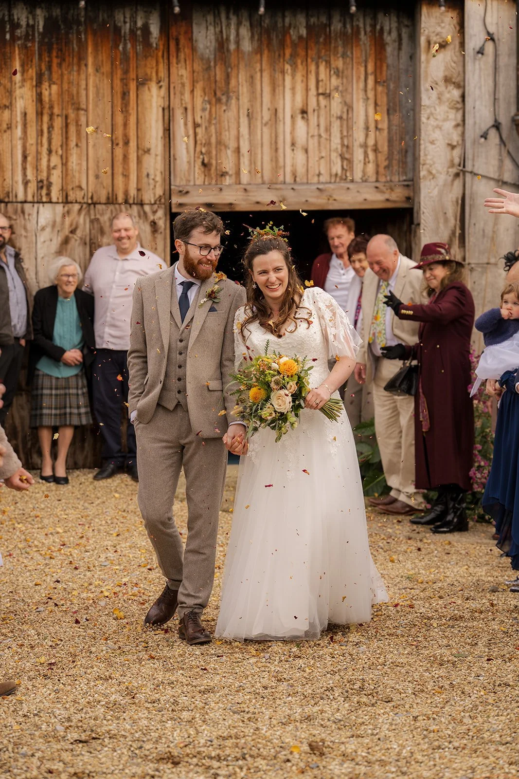 Maisie & Andrew walking through confetti outside Stockbridge Farm Barn in Dorset, smiling together after their autumn ceremony, photographed by Just James Weddings.