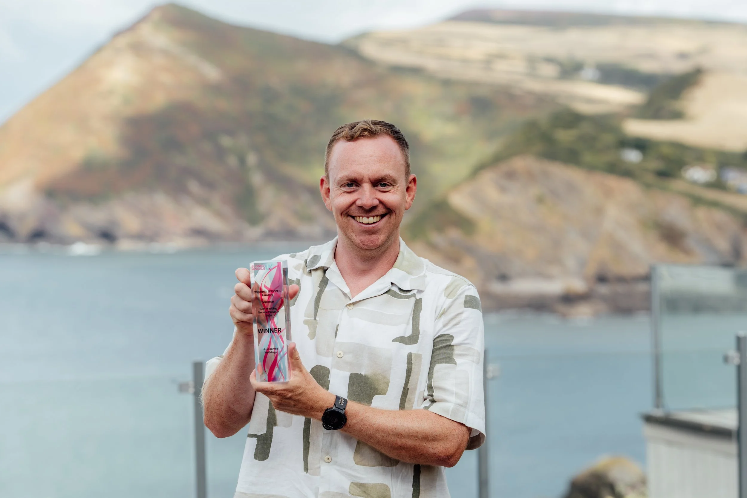 James from Just James Weddings holding his Business Awards UK trophy for winning Wedding Services in Somerset, with the North Devon coastline behind him.
