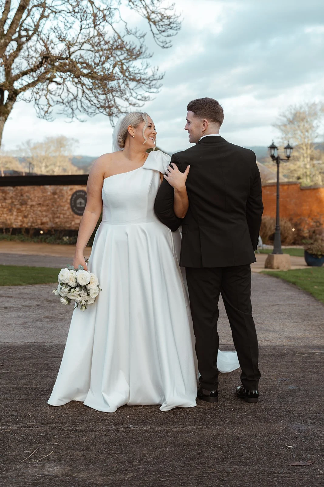 Jessie & Jay walk hand in hand through the grounds of Upton Barn & Walled Garden in Devon, UK. Jessie looks back smiling at Jay as soft winter light and trees frame a relaxed, affectionate moment from their wedding portraits.