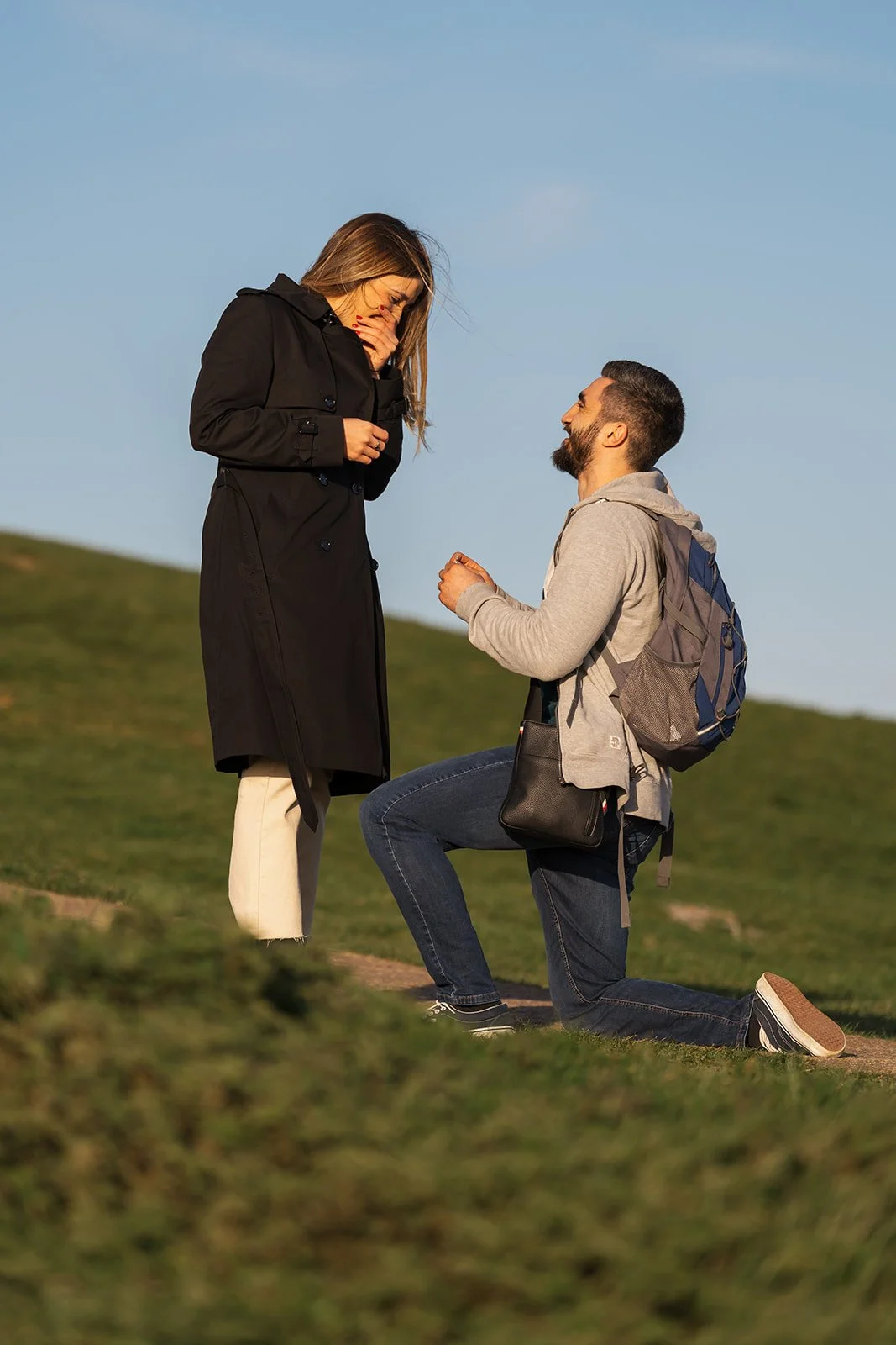 Roy kneeling to propose to Julia on a grassy Somerset hillside, with Julia covering her mouth in surprise against a clear blue afternoon sky during their emotional outdoor engagement photo session, captured by Just James Weddings in Somerset.