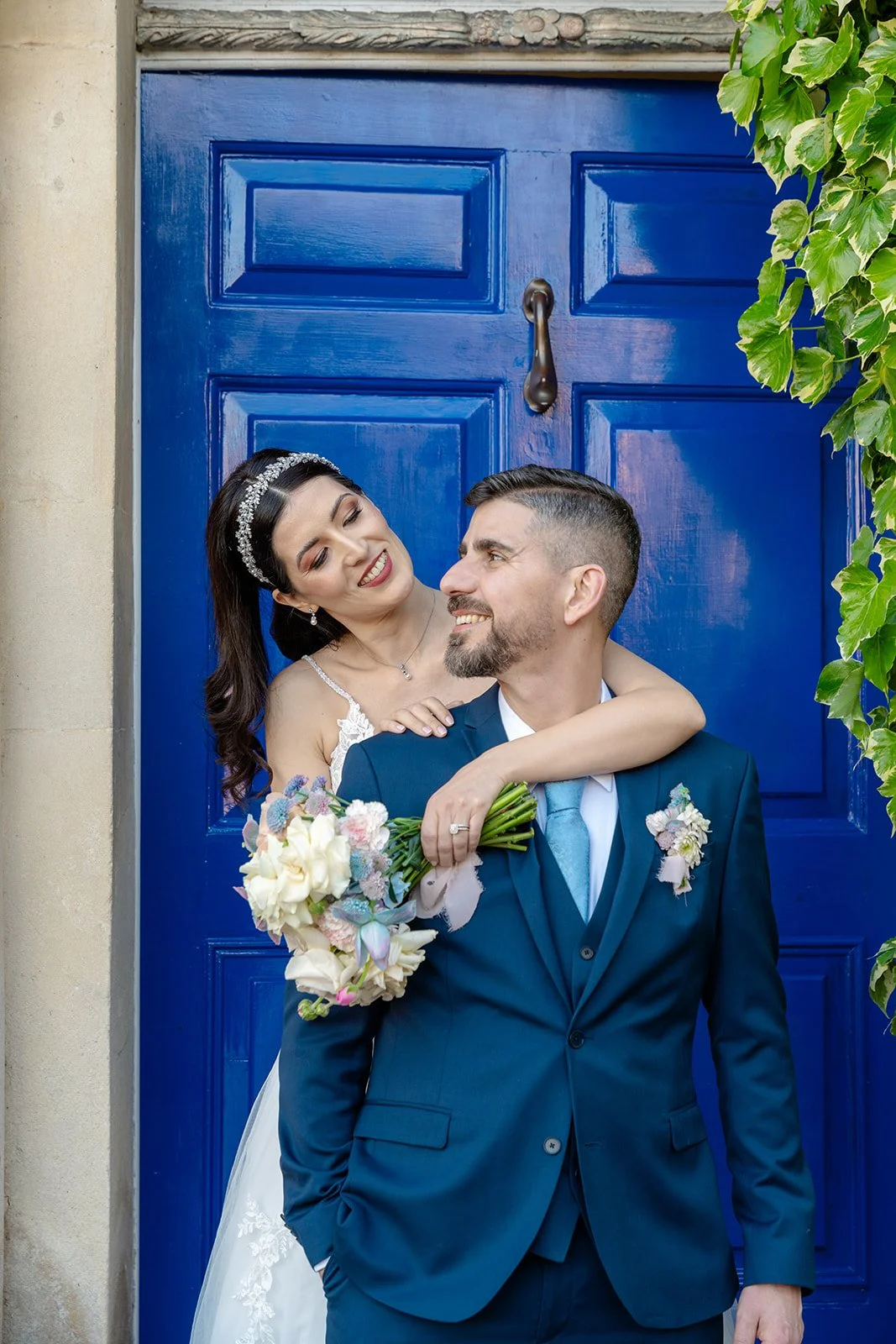 Ruth embraces Carmelo from behind outside No. 4 Clifton Village in Bristol, UK, posing together in front of the venue’s striking blue doorway with her pastel wedding bouquet.