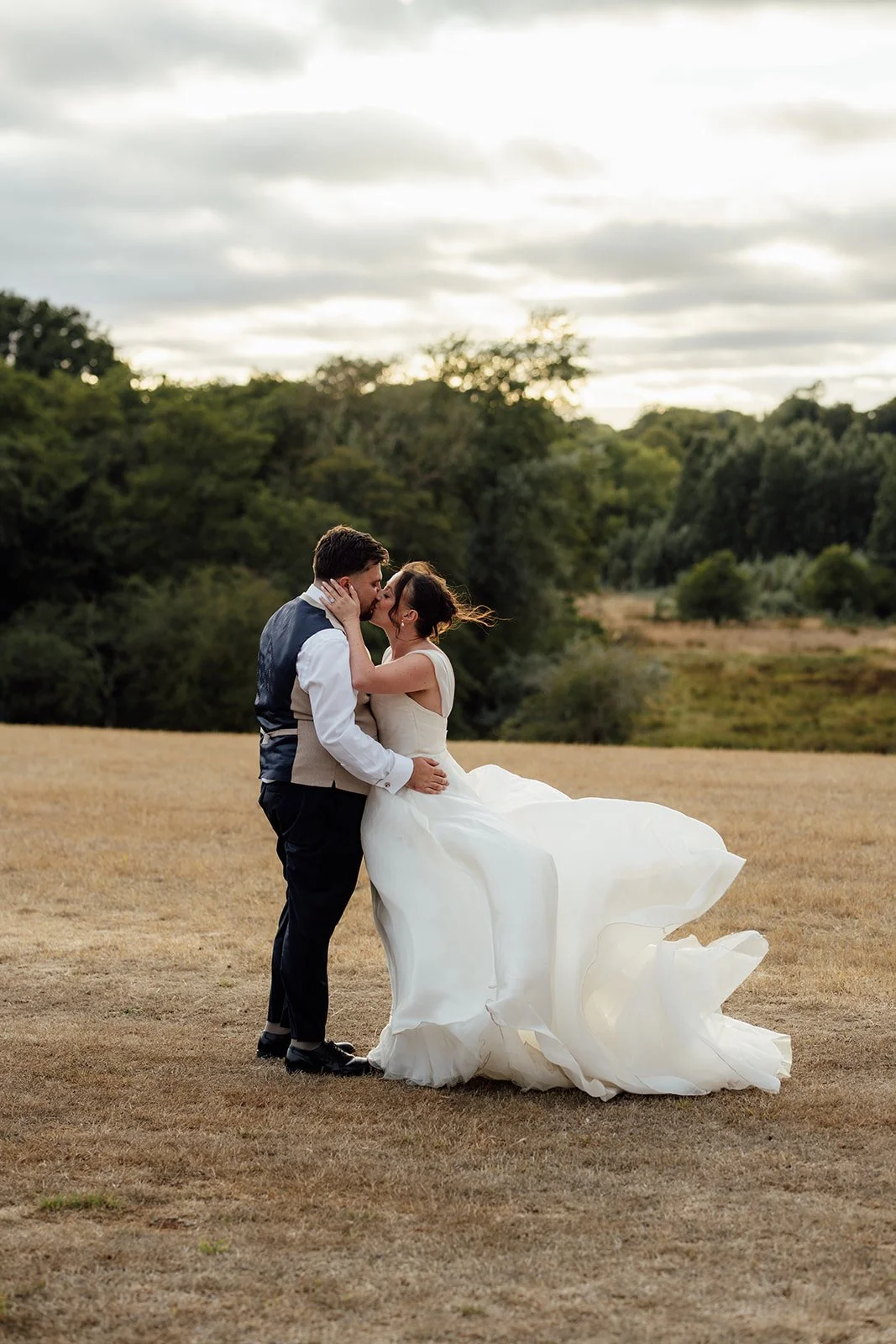 Harriet & Simon share a kiss in the field as Harriet’s wedding dress moves in the breeze at Dovecote Events in Oxfordshire, photographed by Just James Weddings.