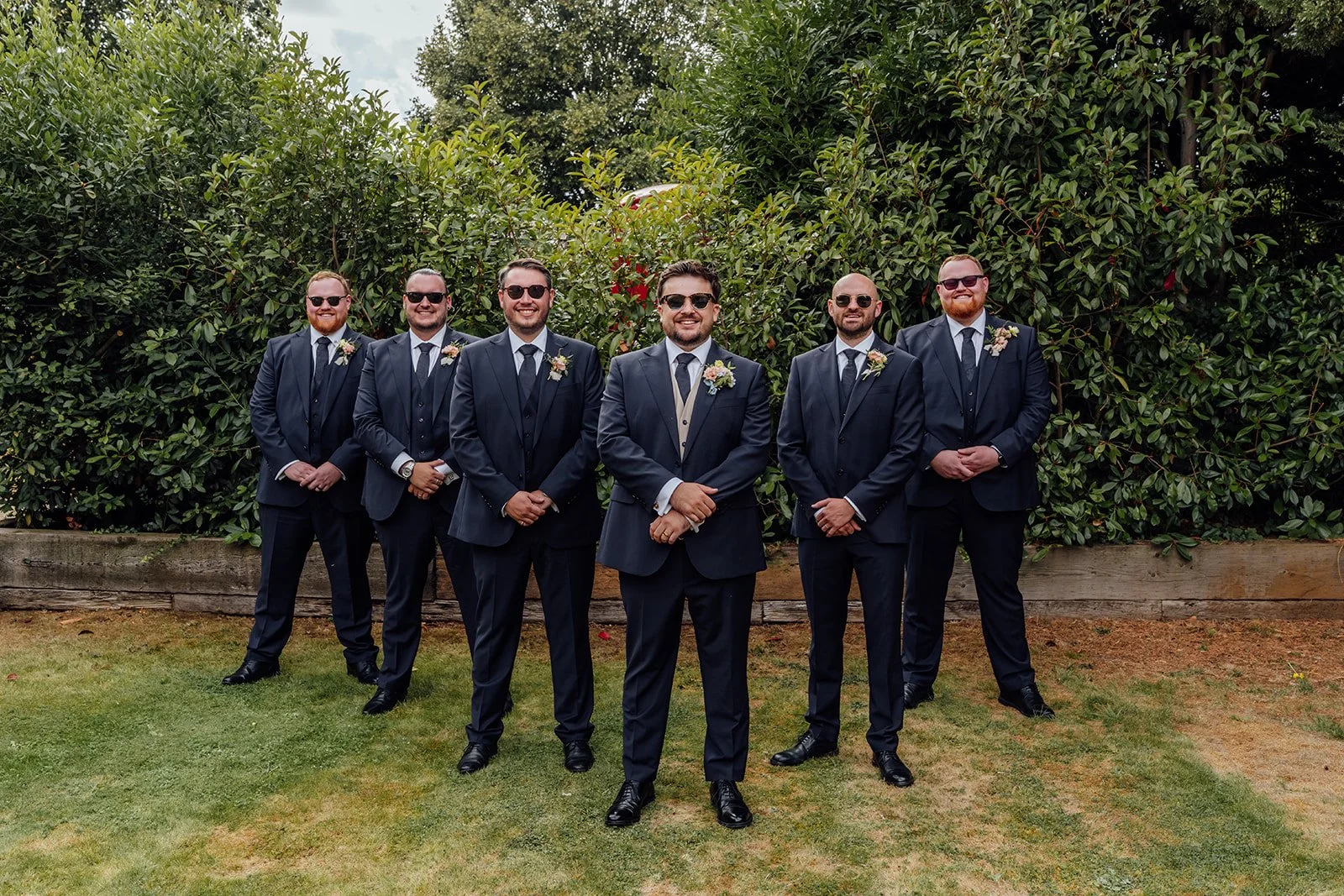 Simon poses with his groomsmen in matching navy suits and sunglasses in the gardens at Dovecote Events in Oxfordshire, photographed by Just James Weddings.