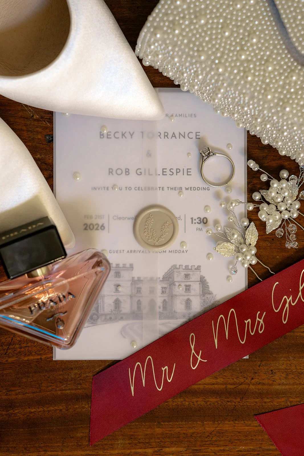 Flat lay of Becky & Rob’s wedding details: Clearwell Castle invitation, engagement ring, pearls, perfume and ivory heels on wood, timeless Clearwell Castle wedding styling in Gloucestershire, UK, for wedding photography in the bridal suite.