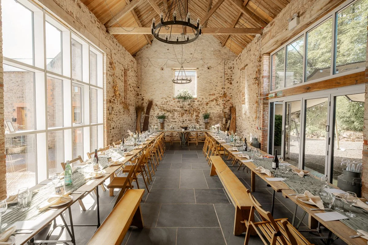 The reception space at The Hawke Barn, devon, styled for Samantha & Mike’s wedding with long wooden tables, bench seating, stone walls, and hanging chandeliers. Soft daylight fills the rustic barn, creating a warm celebration.
