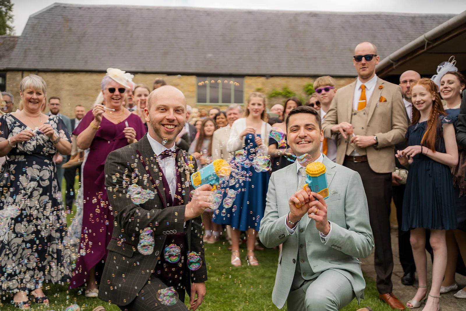Sam & Gavin celebrating with bubbles after their ceremony at The Great Barn at Aynho in the Cotswolds, photographed by Just James Weddings for joyful documentary wedding moments.