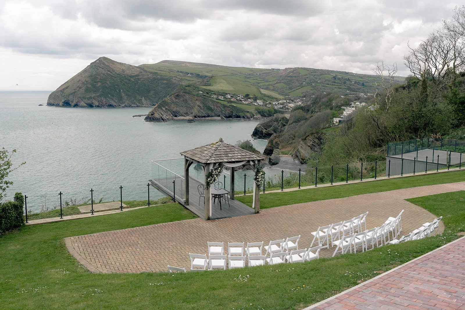 Outdoor ceremony setup at Sandy Cove Hotel in North Devon for Sam & Terri’s wedding, with white chairs, a wooden gazebo and sea views over the cliffs, photographed by Just James Weddings for coastal wedding venue inspiration and wedding ideas.
