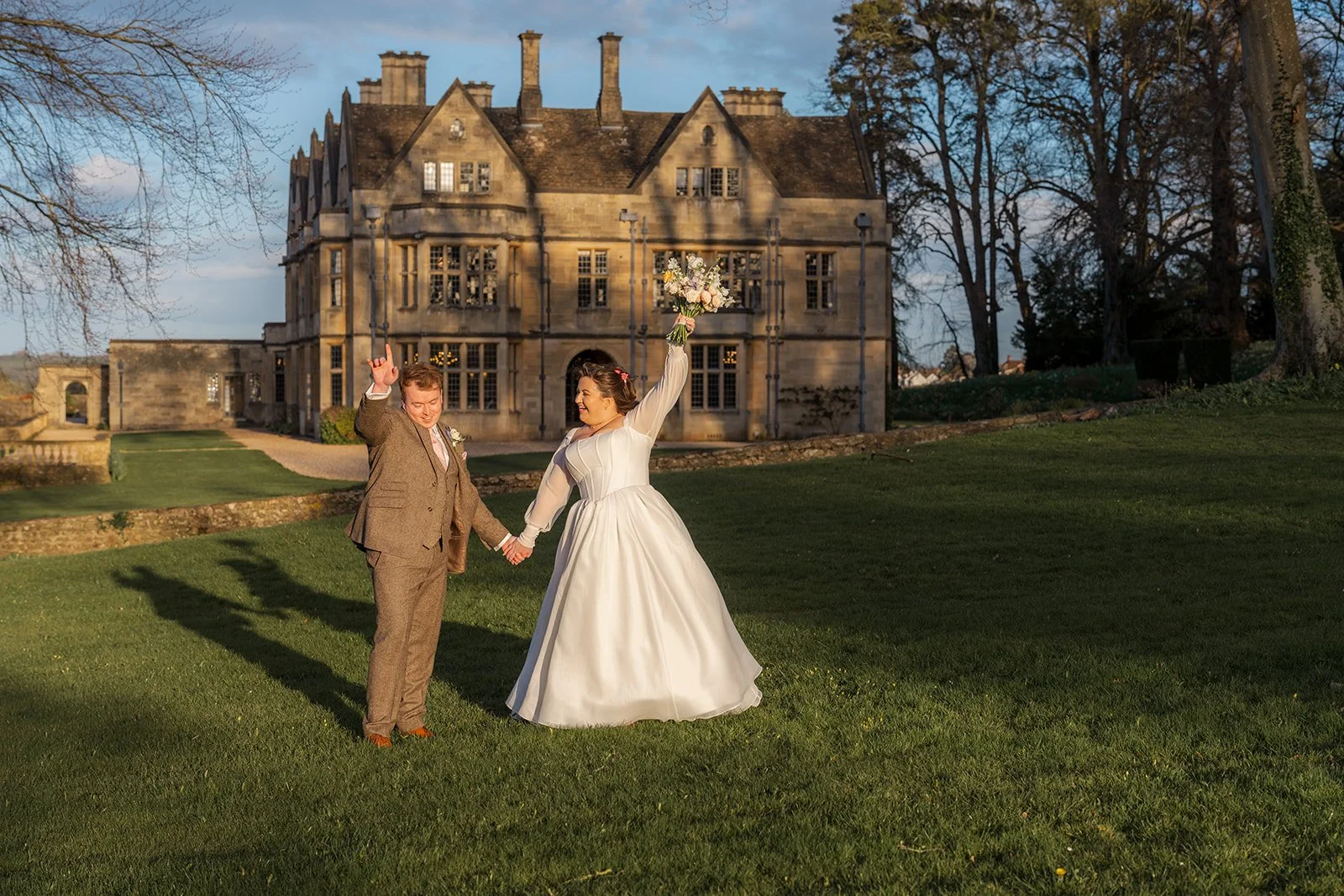 Emily & Josh stand hand in hand on the lawns at Coombe Lodge Blagdon, Somerset, UK, with the historic house behind them in golden evening light. Emily raises her bouquet while Josh lifts his hand, framed by the Somerset venue and wide green grounds.