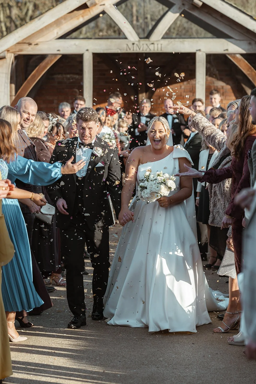 Jessie & Jay walk hand in hand through a joyful confetti exit at Upton Barn & Walled Garden in Devon, UK. Guests line the pathway cheering as petals fall around the couple, capturing laughter, movement, and celebration after the ceremony.