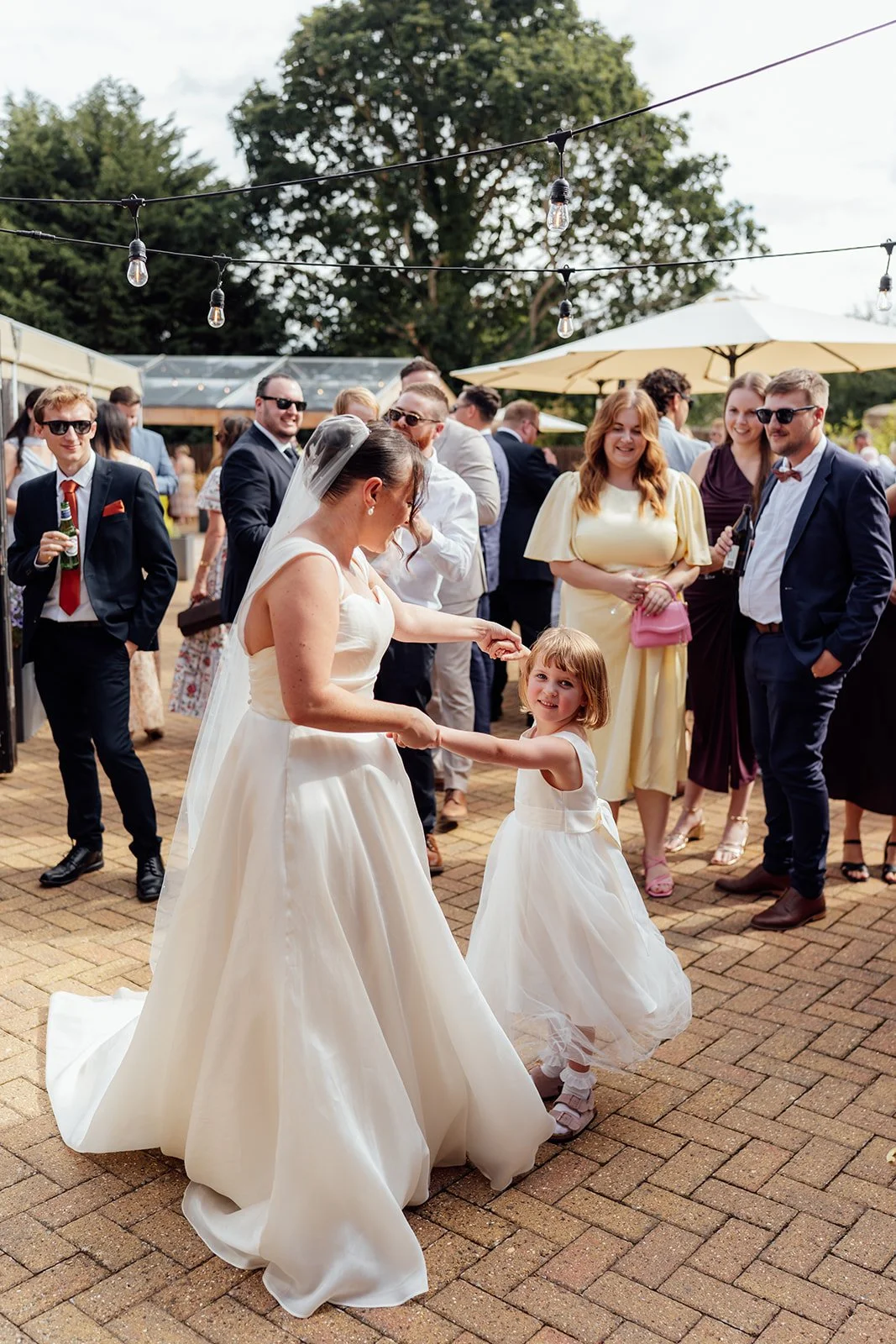 Harriet dances hand in hand with a young flower girl while wedding guests watch beneath festoon lights at Dovecote Events in Oxfordshire, photographed by Just James Weddings.