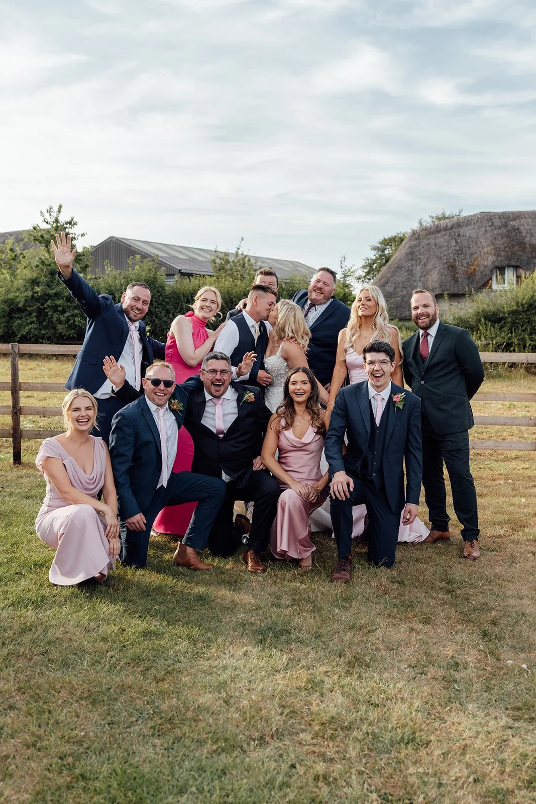 Elise and Tom kissing in the centre of their wedding party during a relaxed countryside group photo at Bowerchalke Barn in Wiltshire, photographed by Just James Weddings.
