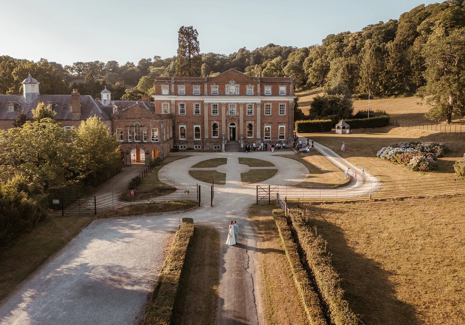 Michelle & Andy stand together for an aerial wedding portrait at Crowcombe Court in Somerset, UK, captured in front of the grand country house with sweeping countryside views, formal gardens and golden evening light.