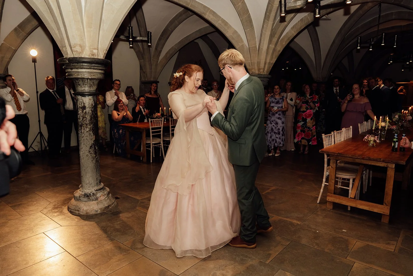 Charlotte & Jack share their first dance at Bishop’s Palace in Somerset, UK, beneath vaulted stone arches as wedding guests watch, cheer and film around candlelit tables during the evening reception inside the historic celebration space nearby.