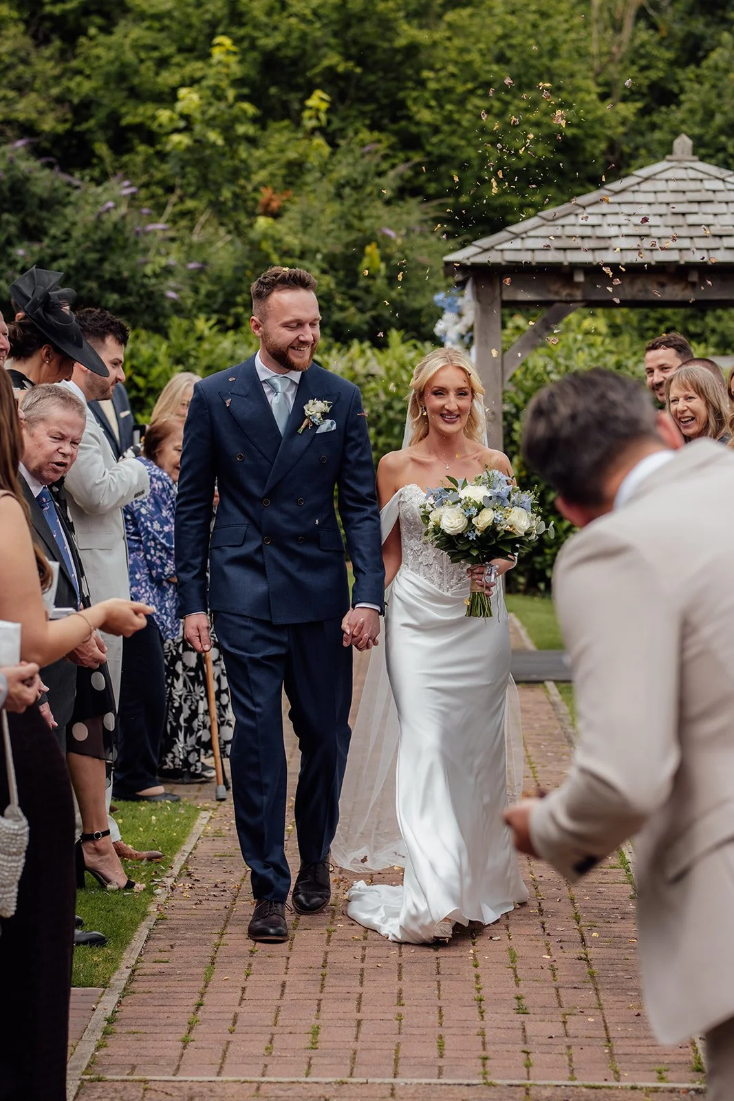 Newlyweds Amy & Connor walk hand in hand through falling confetti after their outdoor wedding ceremony at Sandy Cove Hotel in North Devon, UK.