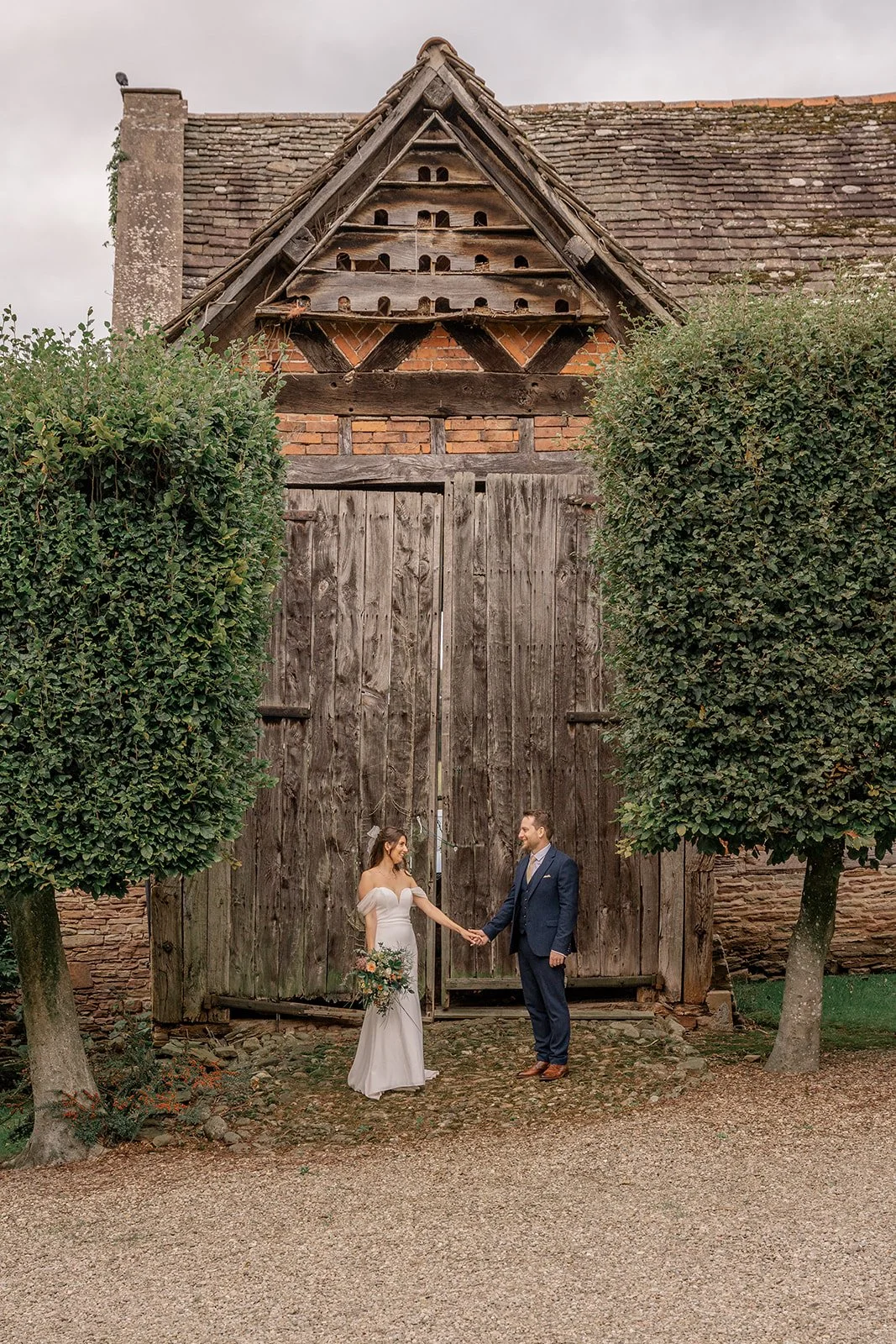 Lilly & Lloyd standing hand in hand in front of a large rustic barn door at Broadfield Court in Herefordshire, photographed by Just James Weddings for natural wedding portraits.
