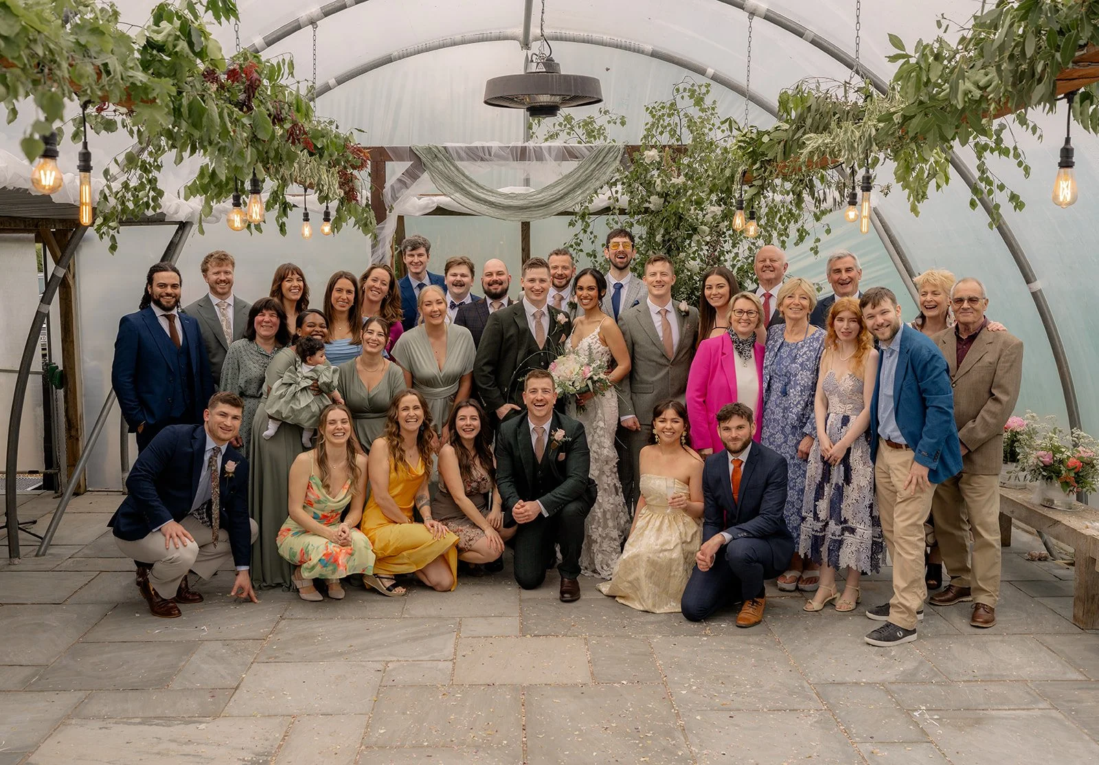Jaye & Josh gather with their guests for a big group photo at Farrington's Farm in Bristol, framed by hanging greenery, festoon bulbs, timber benches, colourful outfits and smiling faces inside the bright poly tunnel wedding ceremony space.