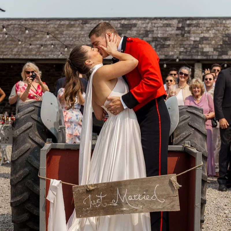 Alba & Josh kissing on the back of a vintage tractor after their wedding ceremony at Bradstone Manor Farm, devon. The guests watch and smile behind them, with rustic stone buildings and gravel courtyard visible.
