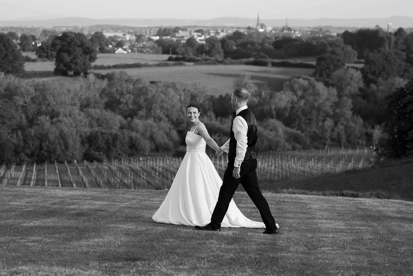 Black and white photo of Lydia and Kieran walking hand in hand with vineyard views during their Hencote Estate wedding in Shropshire, photographed by Just James Weddings.