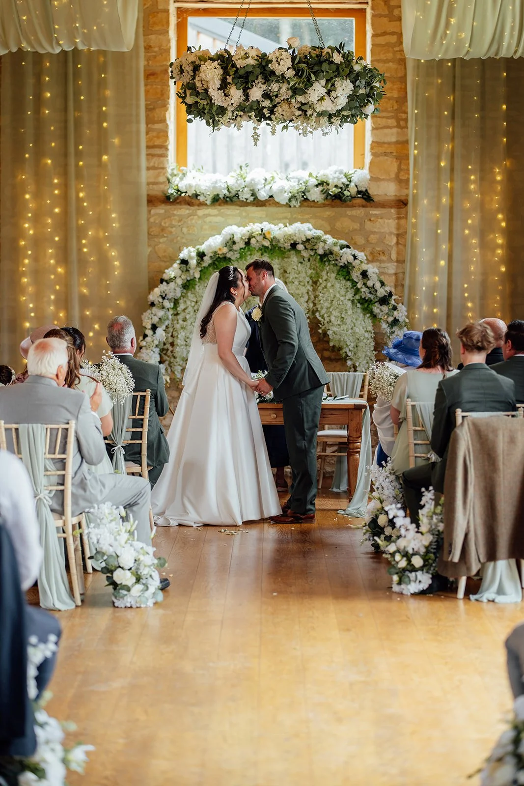 Chloe and Armani share their first kiss during their flower-filled wedding ceremony at The Barn at Upcote in Gloucestershire, photographed by Just James Weddings.
