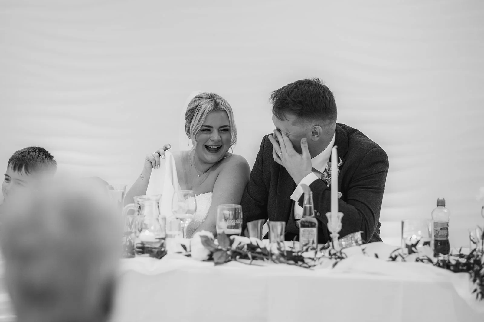 Daisy & Andrew laughing during their wedding speeches at Sandy Cove Hotel in North Devon, captured in black and white by Just James Weddings for candid reception photography with emotional moments, table details and a relaxed guest atmosphere.
