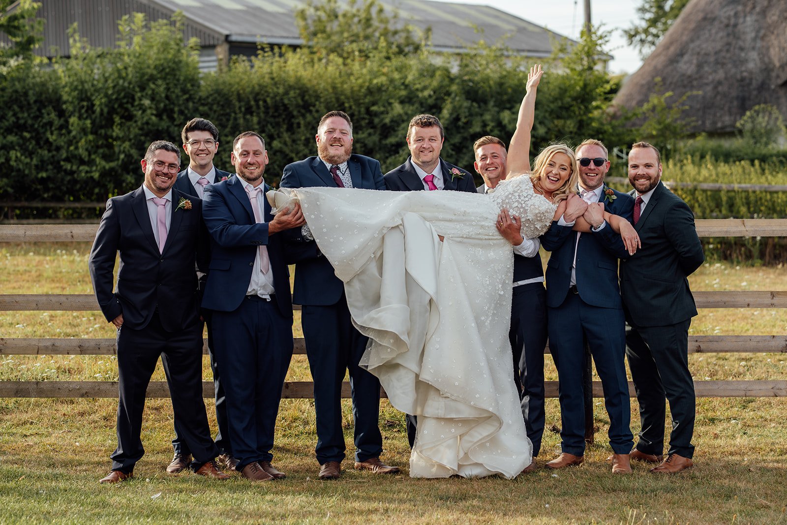 Elise laughing as Tom and his groomsmen lift her during a fun wedding party portrait at Bowerchalke Barn in Wiltshire, photographed by Just James Weddings.