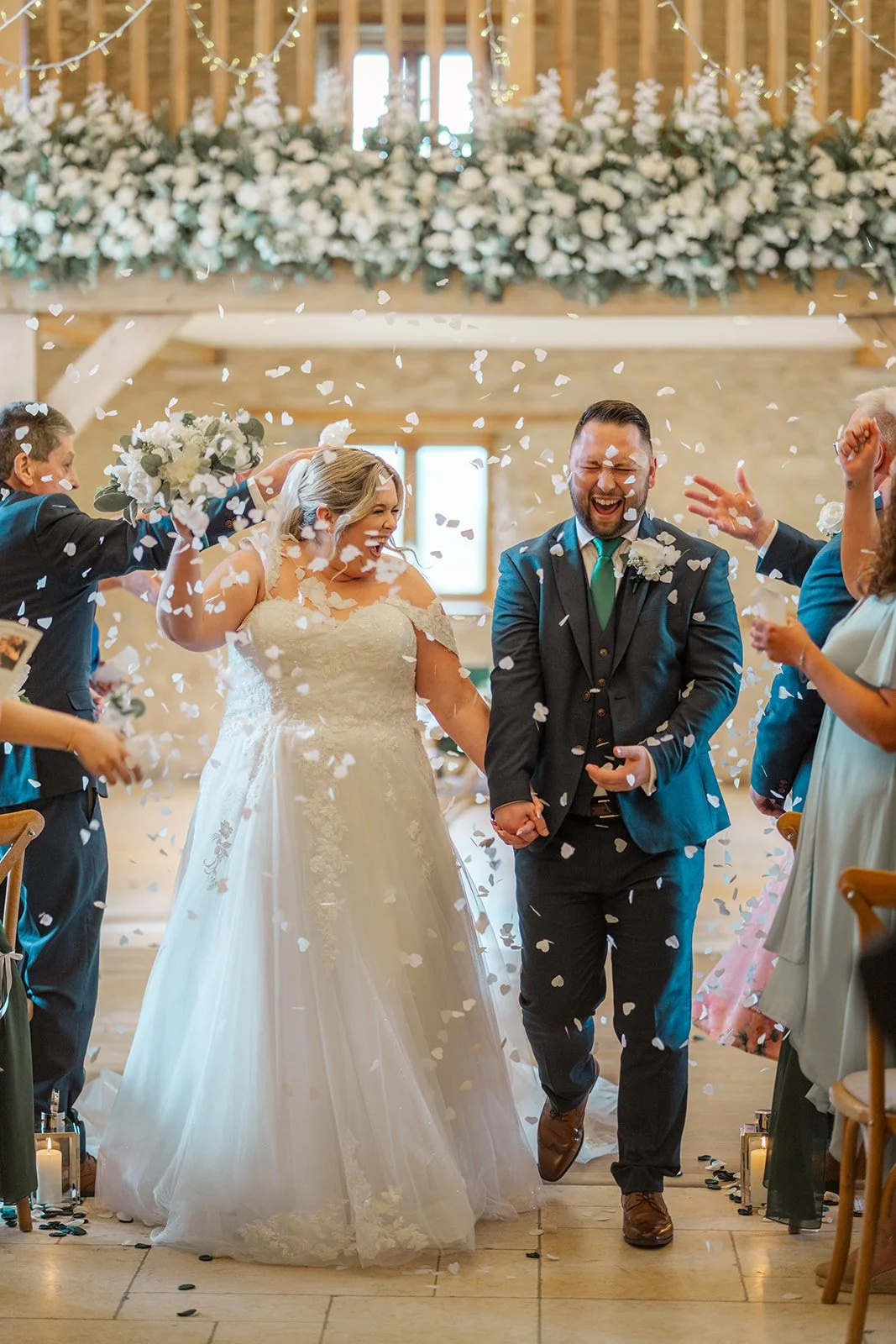 Siobhan & Nicholas walk back down the aisle through a shower of white confetti at Kingscote Barn in the Cotswolds, UK, smiling and holding hands during their rustic barn wedding ceremony exit as guests cheer and celebrate around them indoors.