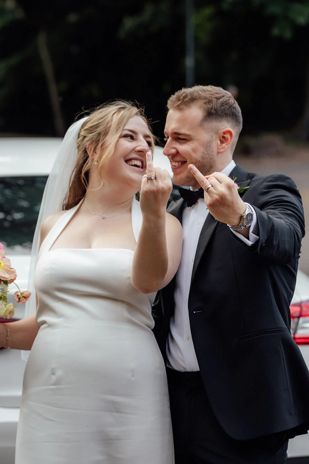 Bryony & Max laugh beside their wedding car at Insole Court in Cardiff, holding up their wedding rings with playful smiles, Bryony in her white dress and Max in his black tuxedo for a fun candid newlywed portrait after the ceremony together.
