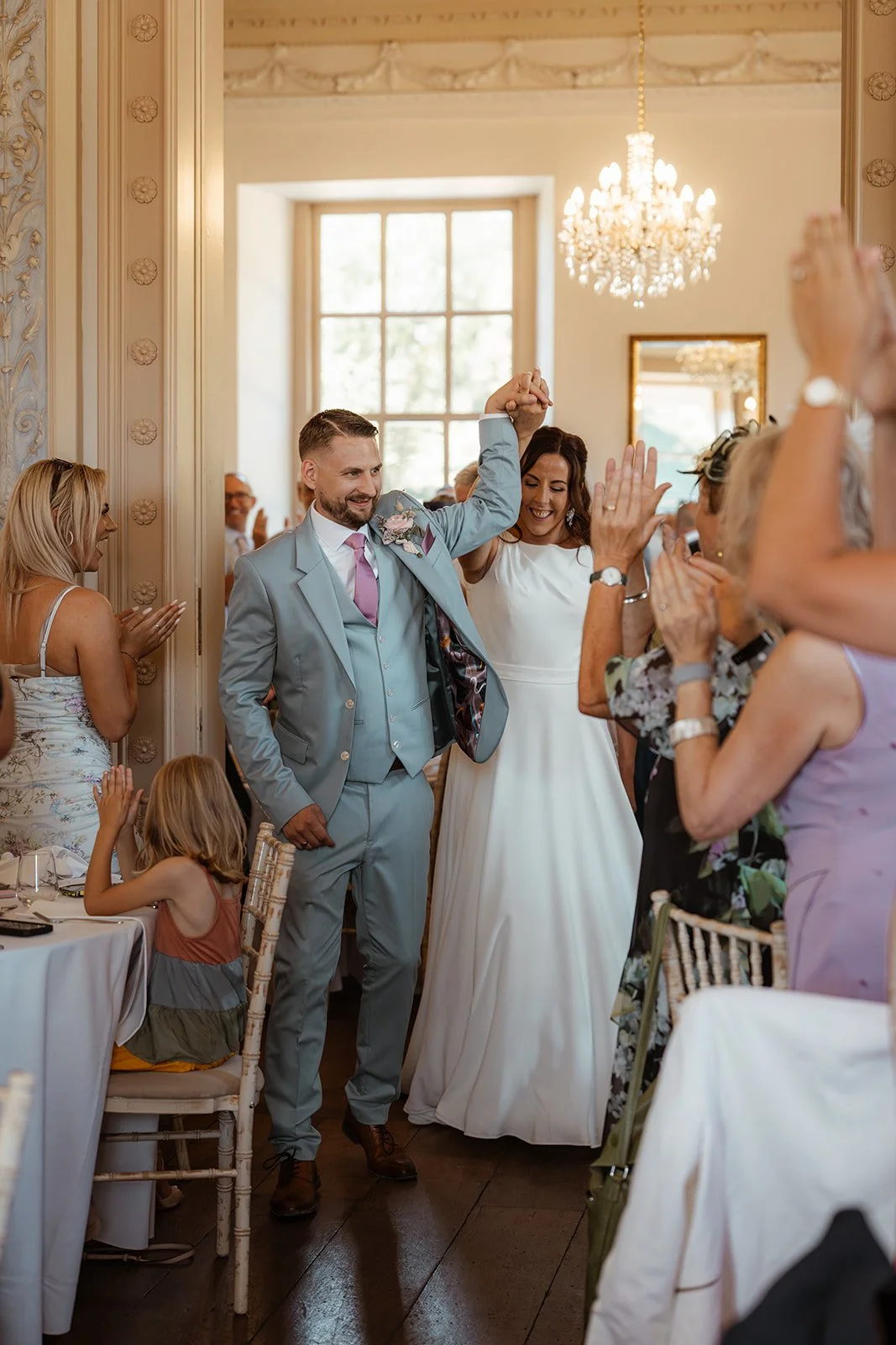 Michelle and Andy make their entrance into their wedding reception at Crowcombe Court in Somerset, UK, holding hands as guests cheer and applaud around them.