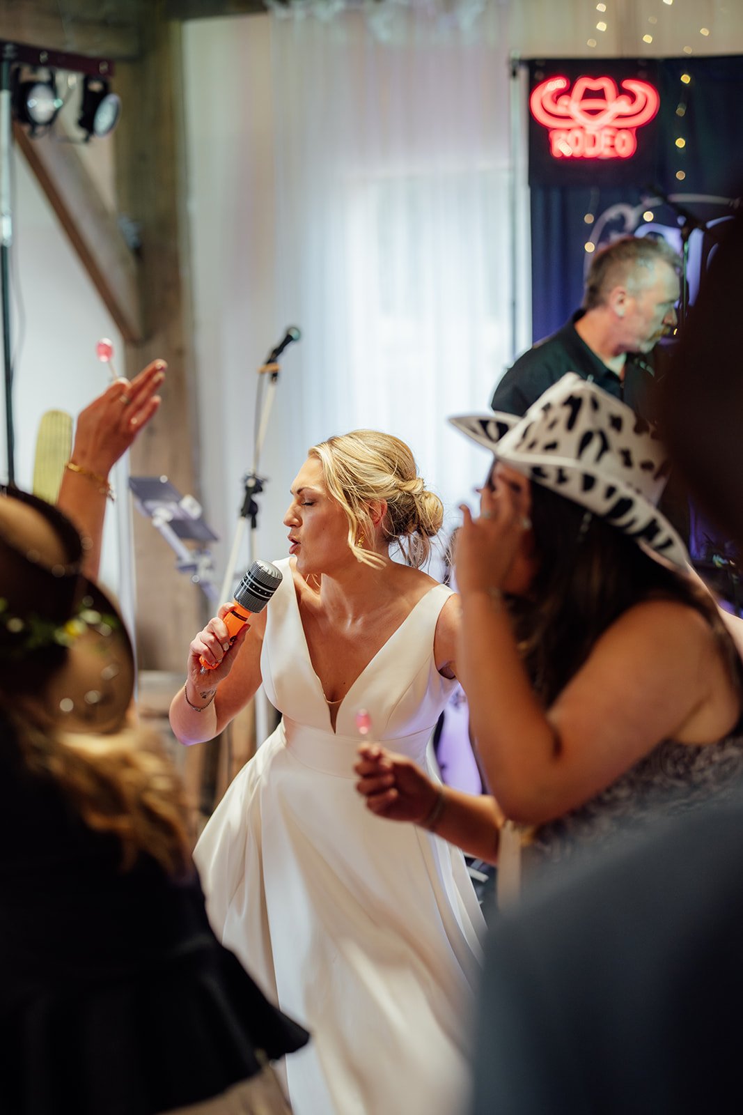 Candid reception photo from Hannah & Ben’s wedding at Grays Barn in Devon, UK, as Hannah sings into a microphone on the dance floor with guests, cowboy hats and live music around her, captured with bright party energy by Just James Weddings.
