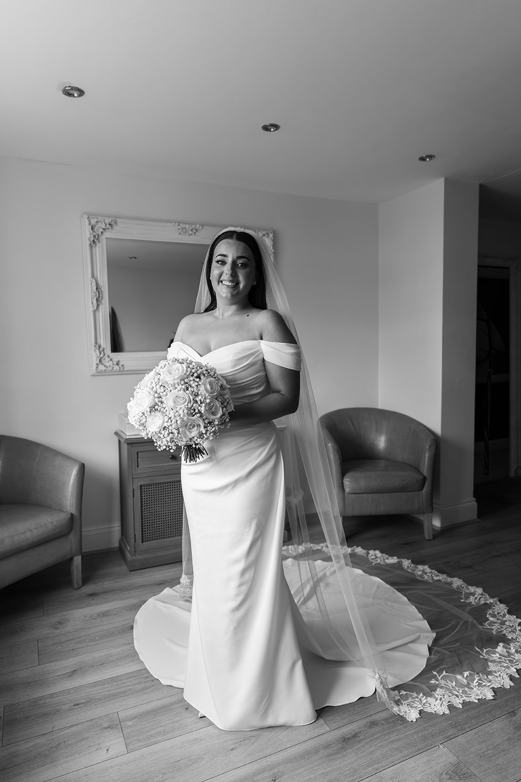 Nisha poses in her wedding dress and veil holding a round bouquet of white roses and baby’s breath before marrying Jack at Batch Country House in Somerset, UK, in the getting-ready room for a bridal portrait, photographed by Just James Weddings.