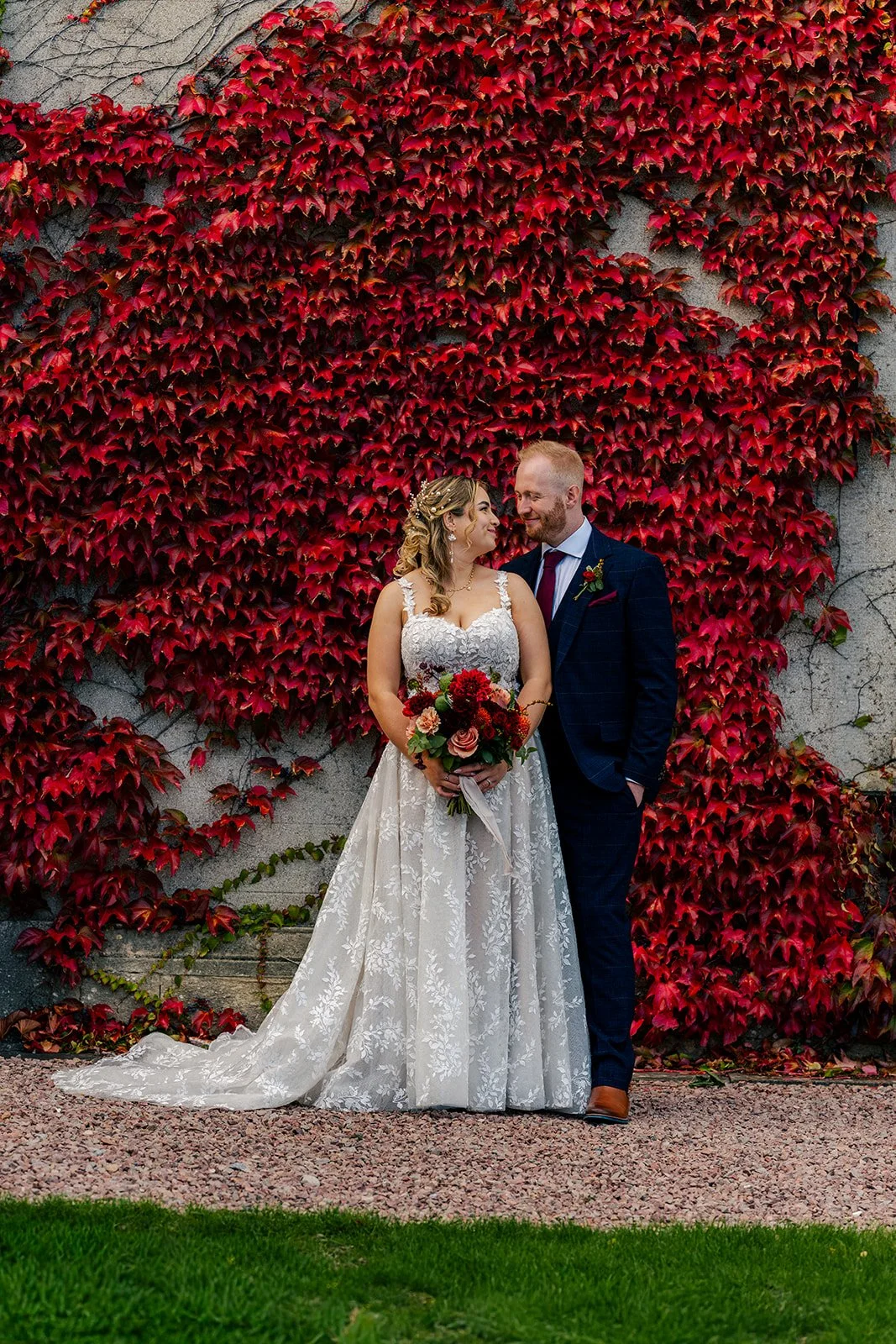 Lucy & Jay standing together in front of red ivy at Eastwood Park in Gloucestershire, photographed by Just James Weddings for colourful autumn wedding portraits.