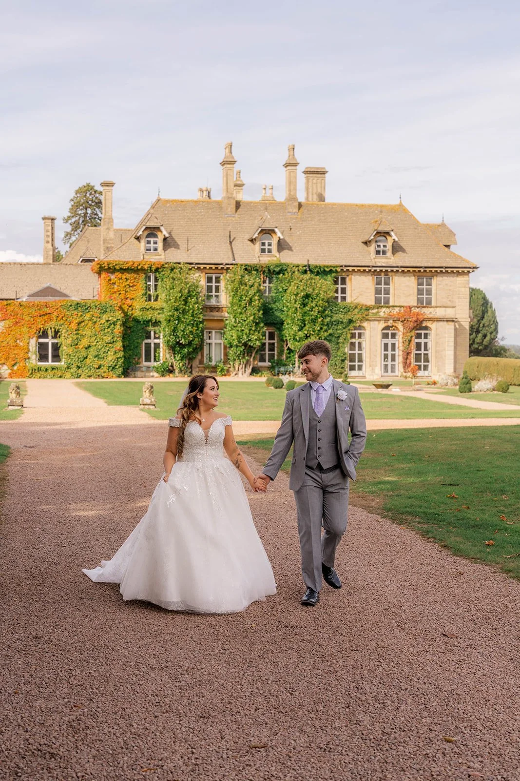 Rhiannon & Anthony walking hand in hand outside Eastwood Park in Gloucestershire, with the manor house behind them, photographed by Just James Weddings for relaxed wedding portraits.