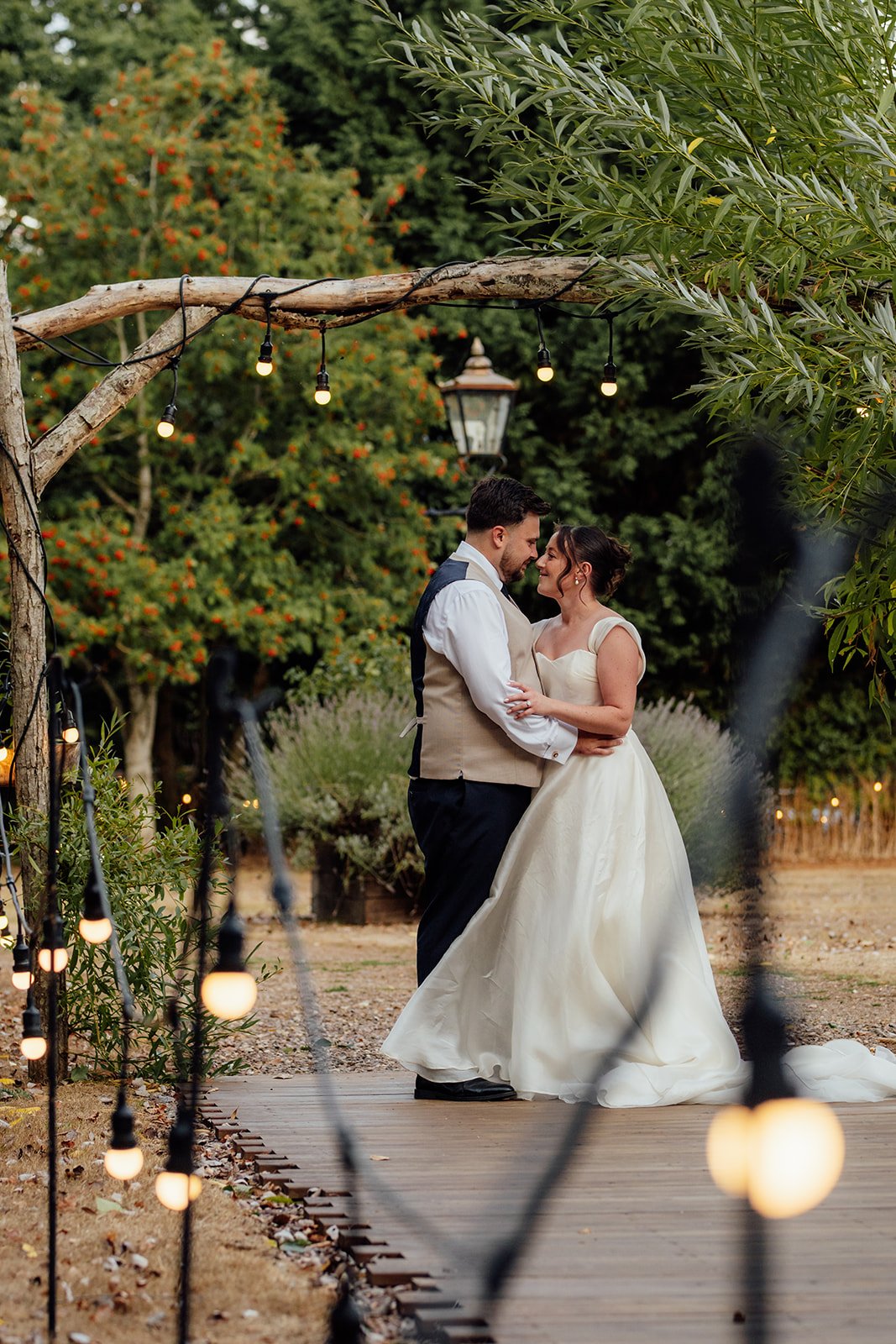 Harriet & Simon embrace beneath festoon lights along a wooden walkway in the gardens at Dovecote Events in Oxfordshire, photographed by Just James Weddings.