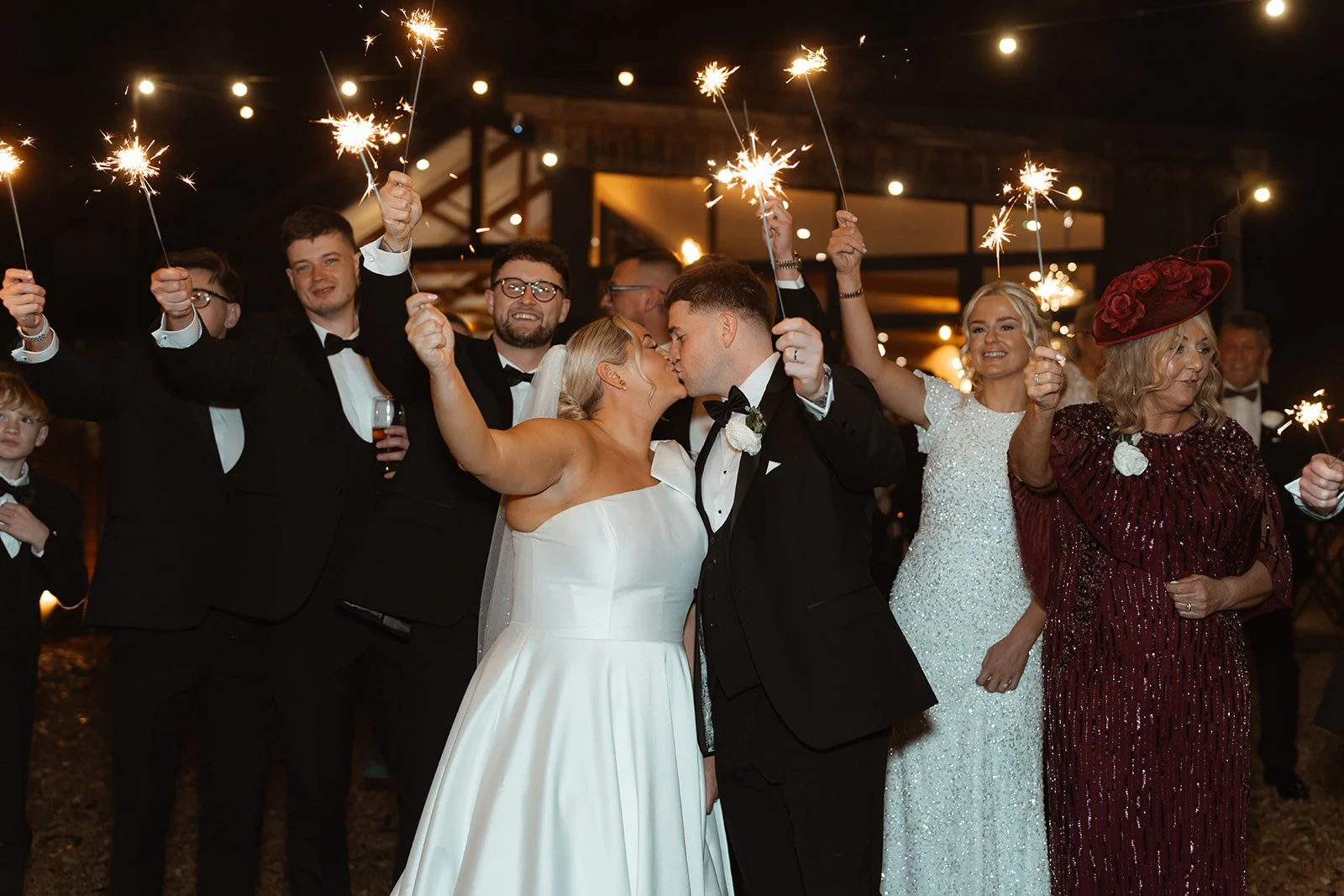 Jessie & Jay share a kiss during a sparkler exit at Upton Barn & Walled Garden in Devon, UK. Friends and family surround the couple holding sparklers aloft, creating a joyful, glowing finale to their wedding day.