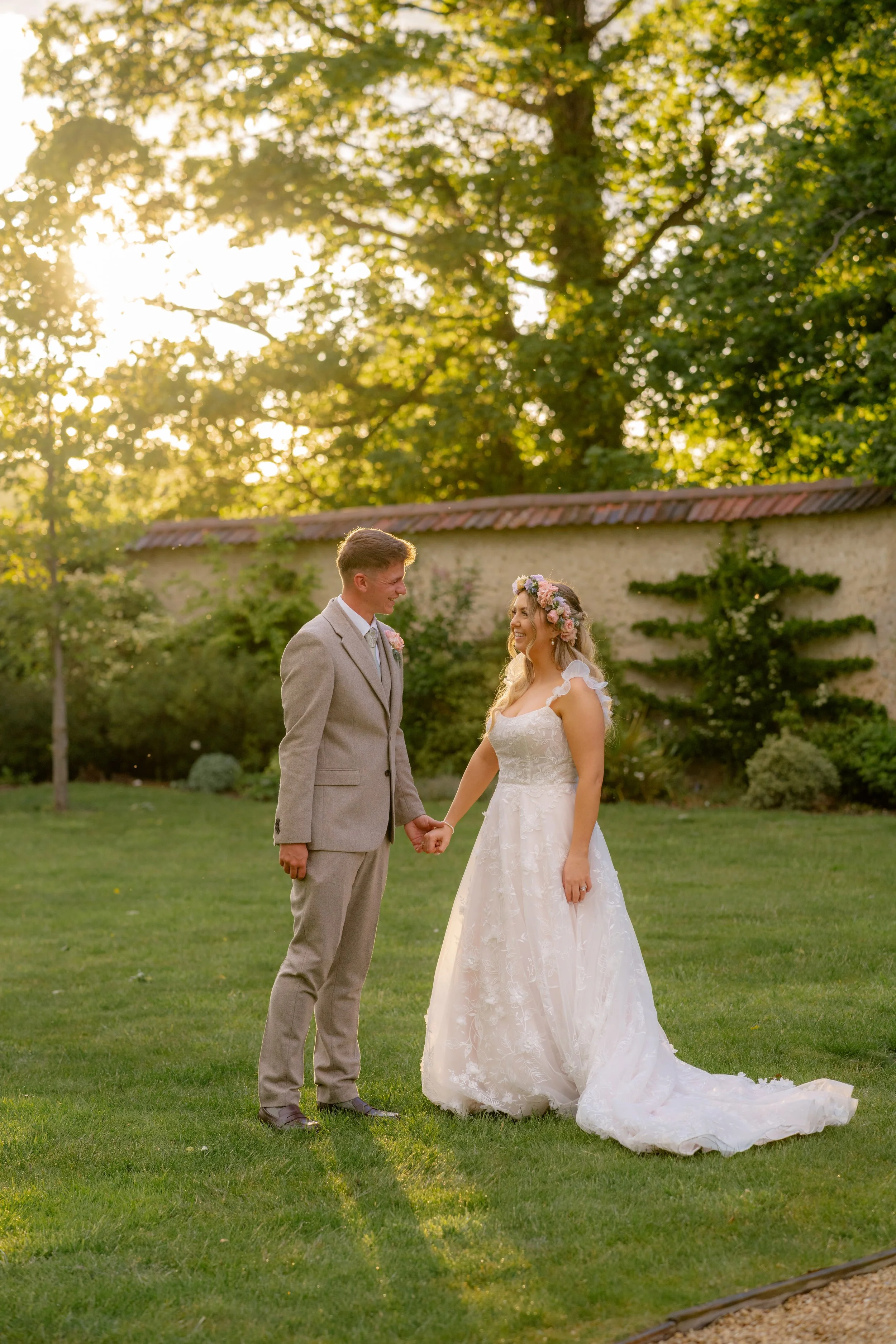 Jaye & Josh’s wedding at Syrencot in Wiltshire, captured in a soft golden hour portrait as the couple stand hand in hand on the lawn, smiling at each other beneath glowing trees, with her floral dress trailing across the grass and warm light.