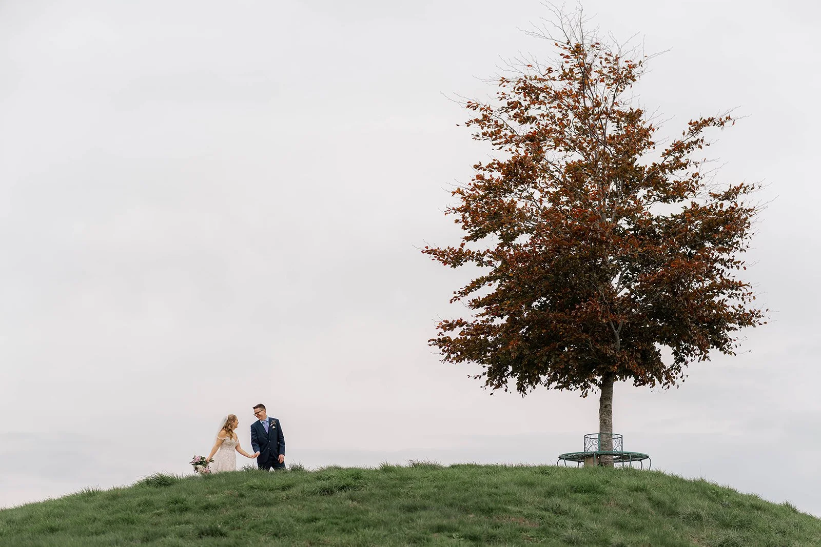 Izzy and Andy walk hand in hand along a grassy ridge, glancing toward each other as they move beneath an open sky. A single tree stands nearby, framing the quiet moment as they stroll together during their wedding portraits.