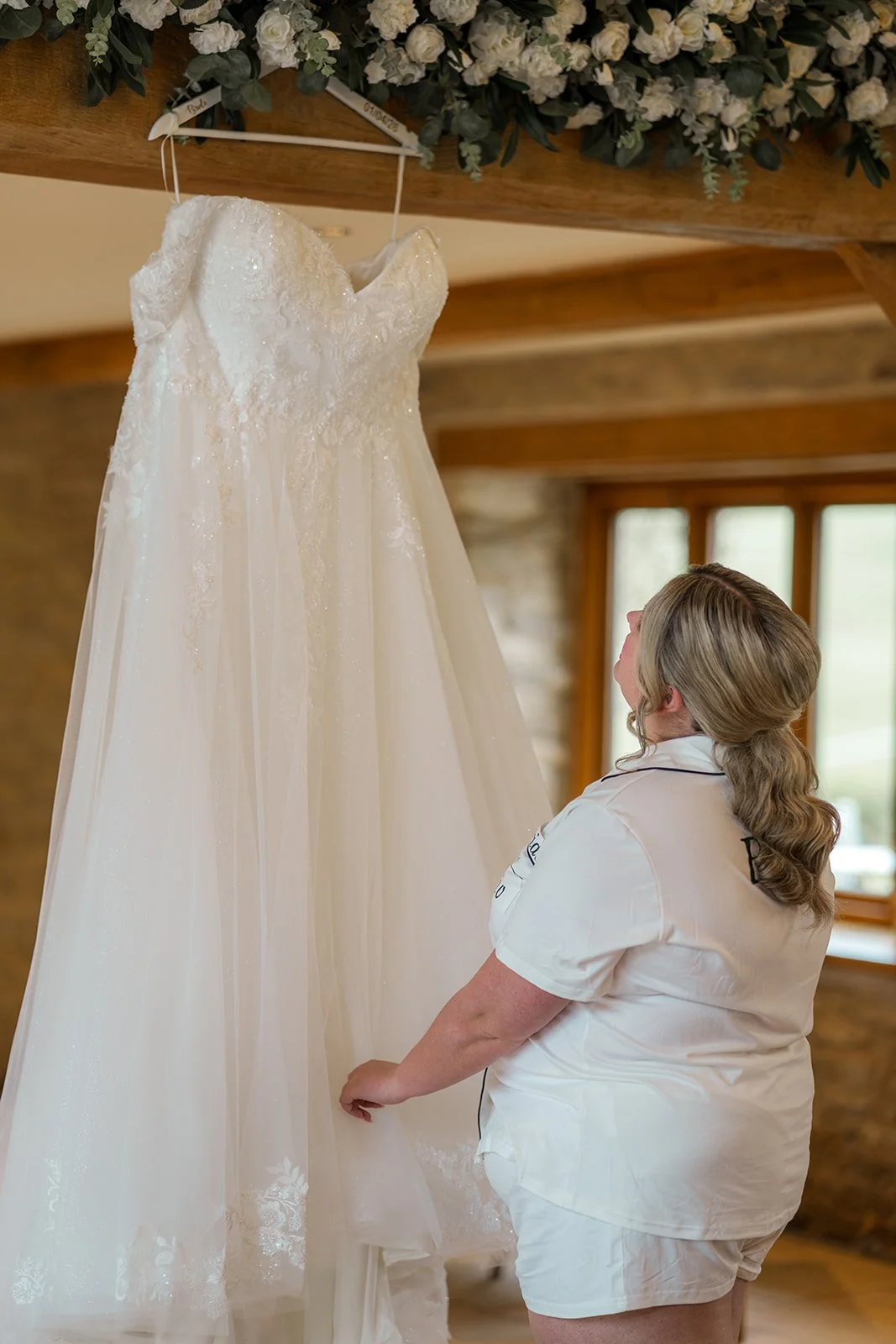 Bride in white pyjamas admiring her lace wedding dress hanging beneath florals inside Kingscote Barn at Siobhan & Nicholas' wedding in the Cotswolds, UK, a soft bridal prep moment in a rustic countryside barn wedding venue with morning light.